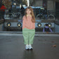 A young girl stands in front of a Paris Metro station entrance, wearing a pink long-sleeve top, light green trousers, and white sneakers.