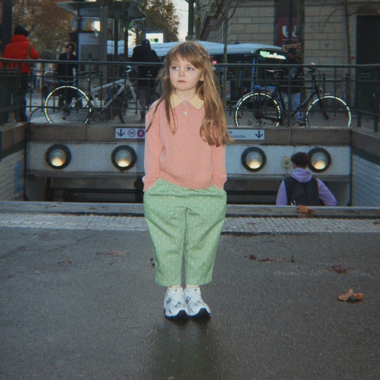 A young girl stands in front of a Paris Metro station entrance, wearing a pink long-sleeve top, light green trousers, and white sneakers.