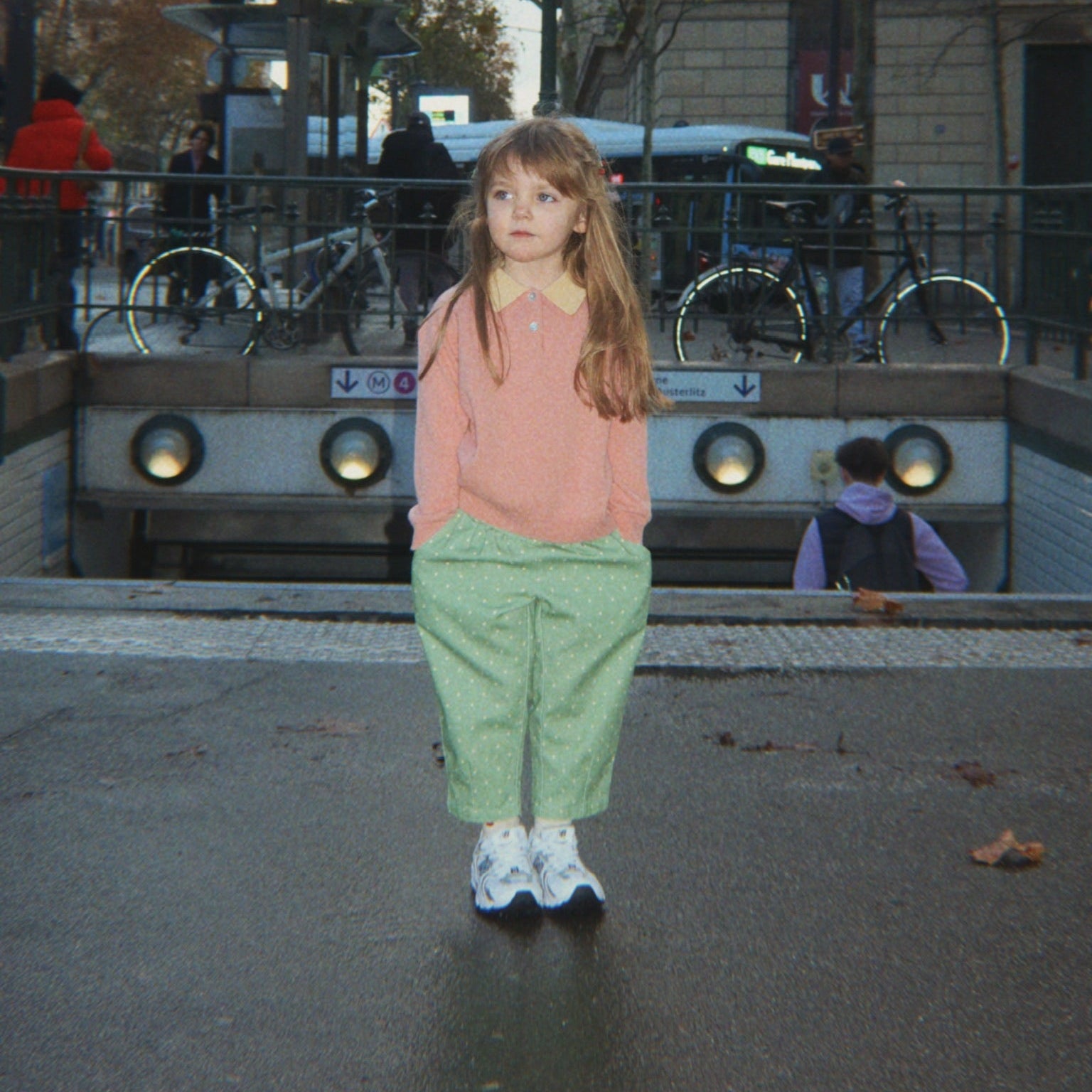 A young girl stands in front of a Paris Metro station entrance, wearing a pink long-sleeve top, light green trousers, and white sneakers.
