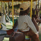 A child wearing a beige baseball cap, a black and white striped long-sleeve shirt, and brown pants sits on a carousel horse in a park.
