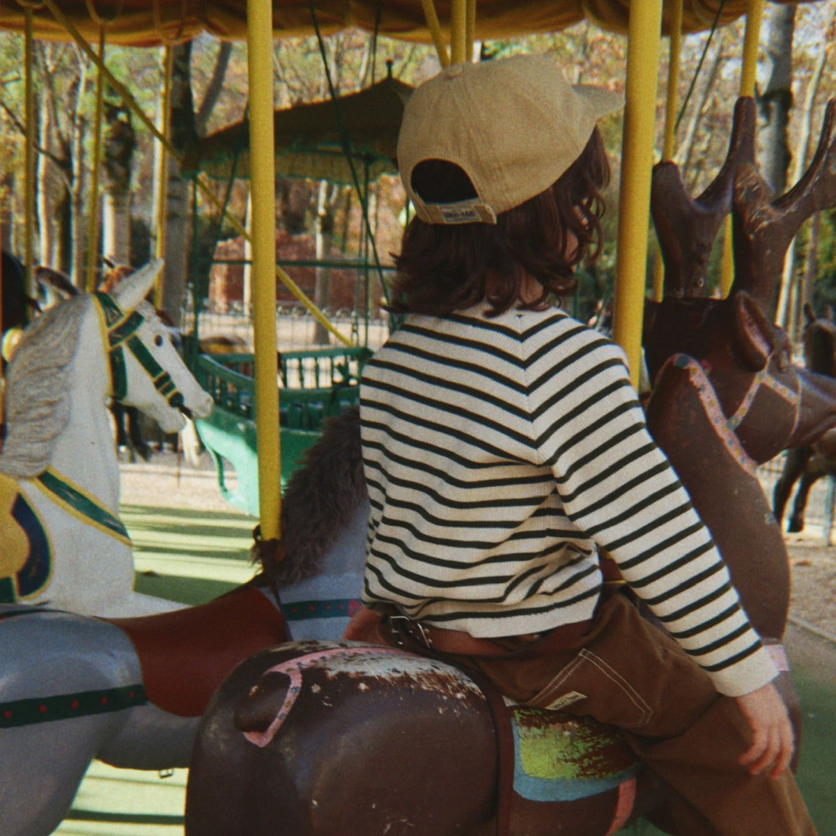 A child wearing a beige baseball cap, a black and white striped long-sleeve shirt, and brown pants sits on a carousel horse in a park.