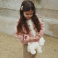 A young girl with long brown hair wearing a pink embroidered jacket, white top, brown checkered pants, and brown Mary Jane shoes, holding a white plush toy against a stone wall background.