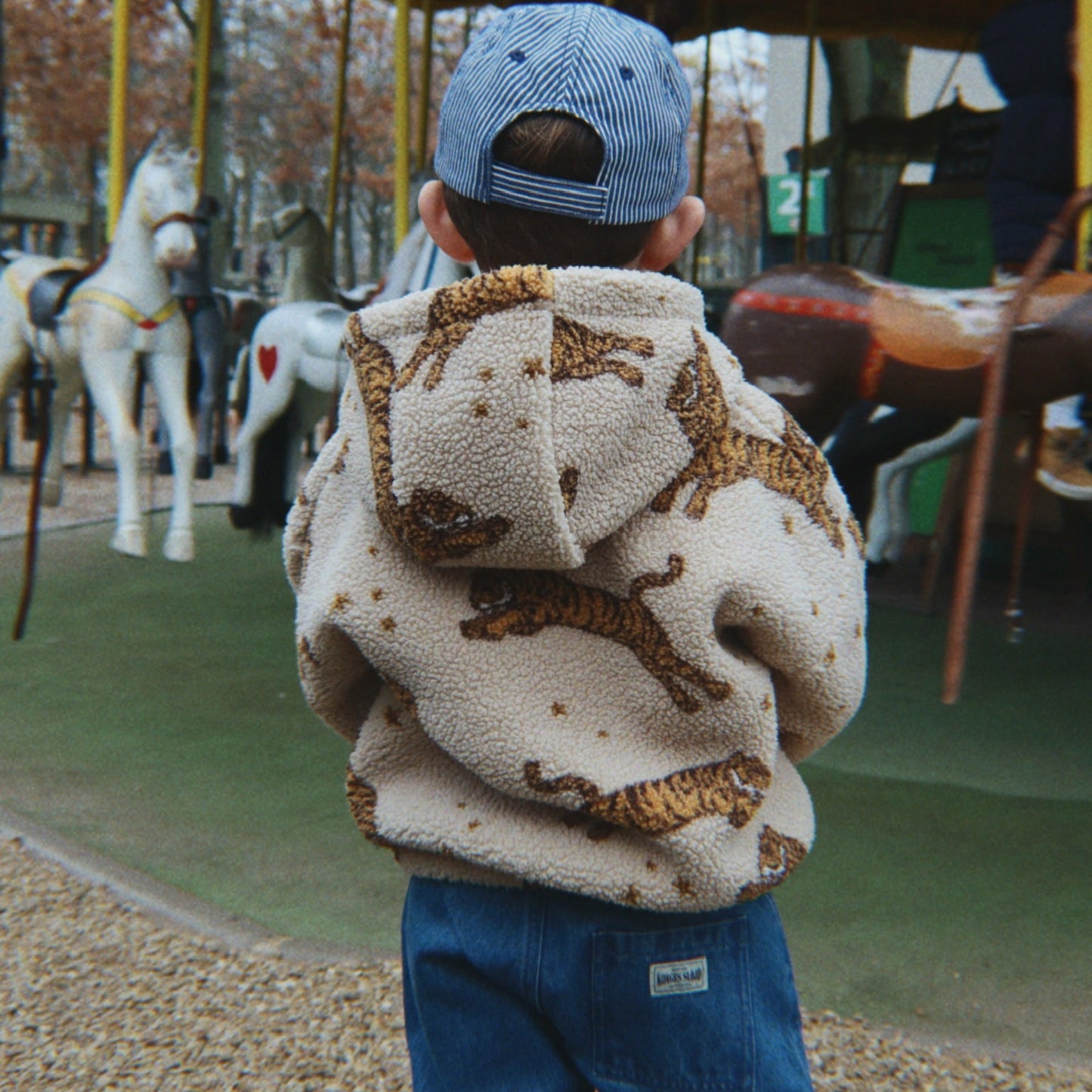A child viewed from behind wearing a cream sherpa hoodie with a brown animal print, blue jeans, and a blue cap, standing in front of a carousel at an outdoor playground.
