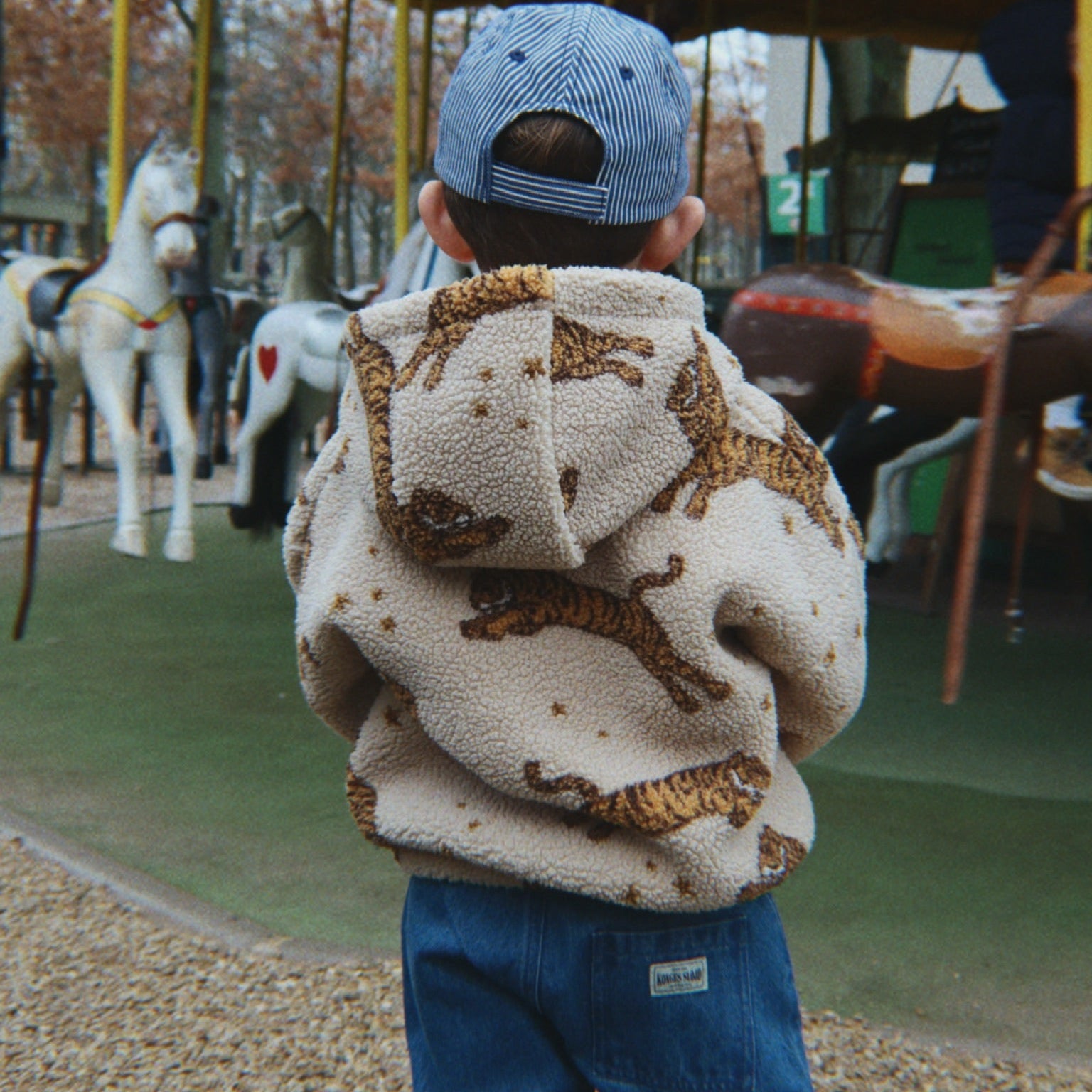A child viewed from behind wearing a cream sherpa hoodie with a brown animal print, blue jeans, and a blue cap, standing in front of a carousel at an outdoor playground.