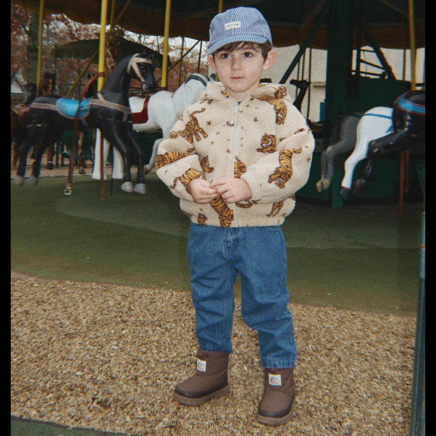 Child wearing a patterned jacket and blue jeans standing on a carousel with horses.