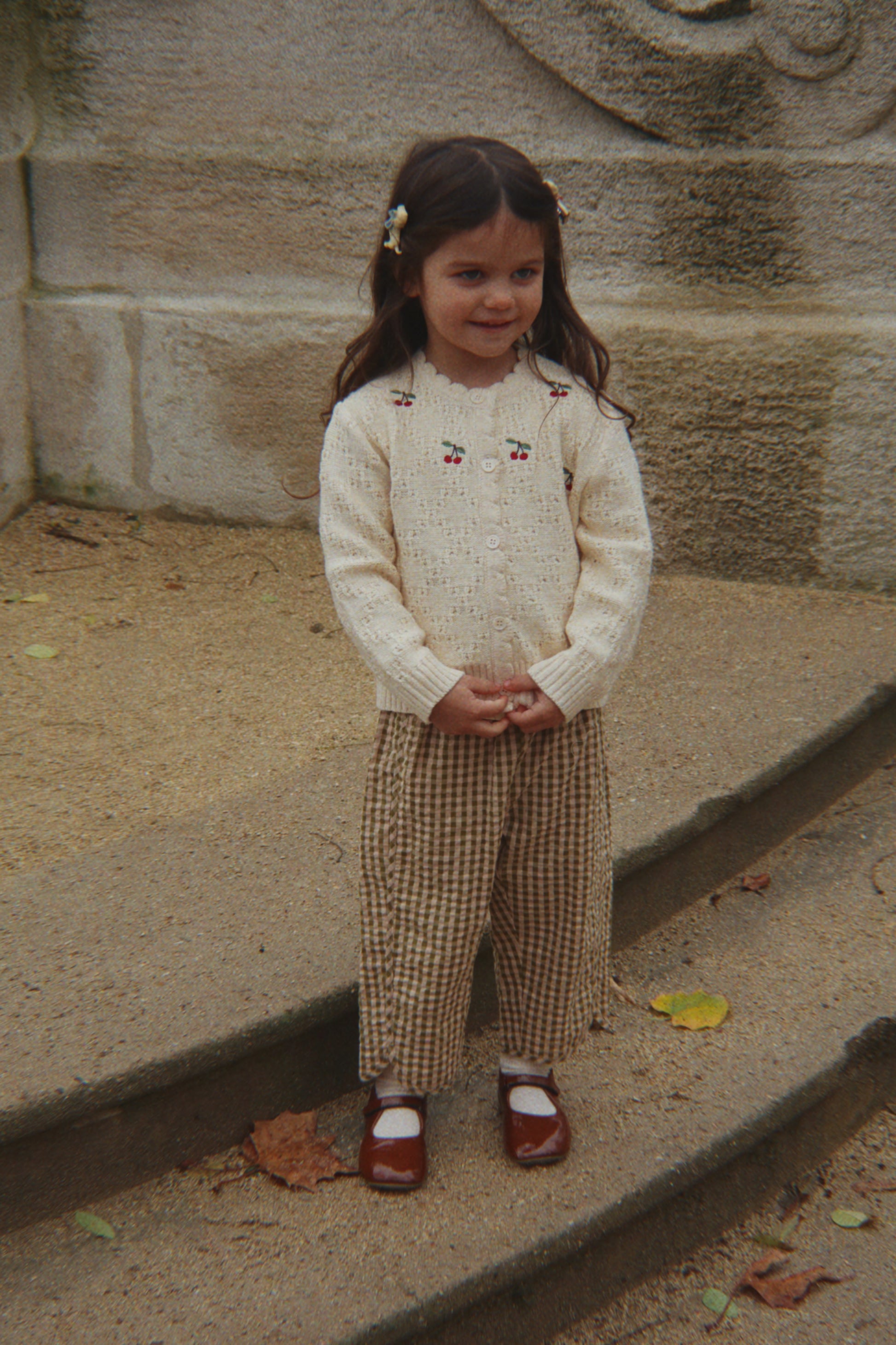 Young girl in a cream sweater and checkered pants standing on steps with a stone wall background