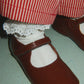 A close-up of a doll's feet wearing shiny dark brown Mary Jane shoes, white lace-trimmed socks, and red-and-white striped pants.