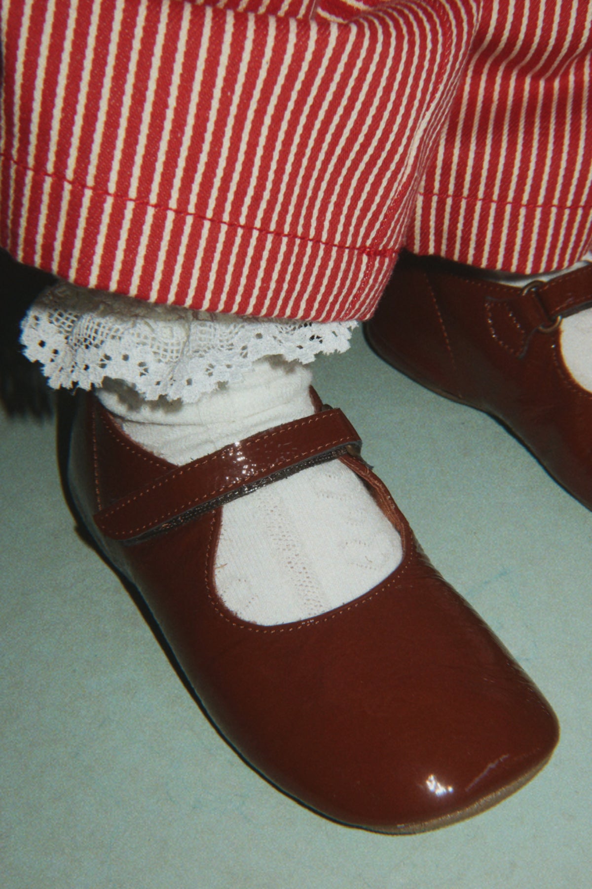 A close-up of a doll's feet wearing shiny dark brown Mary Jane shoes, white lace-trimmed socks, and red-and-white striped pants.
