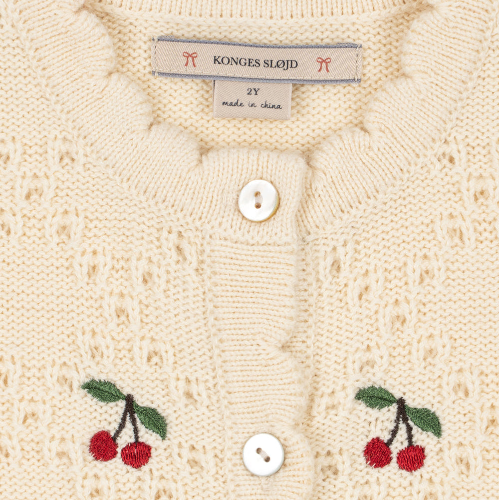 Close-up of a cream-colored knit cardigan featuring scalloped edges, white buttons, and embroidered red cherries with green leaves on both sides. The brand label reads 'KONGJES SLAJID'.