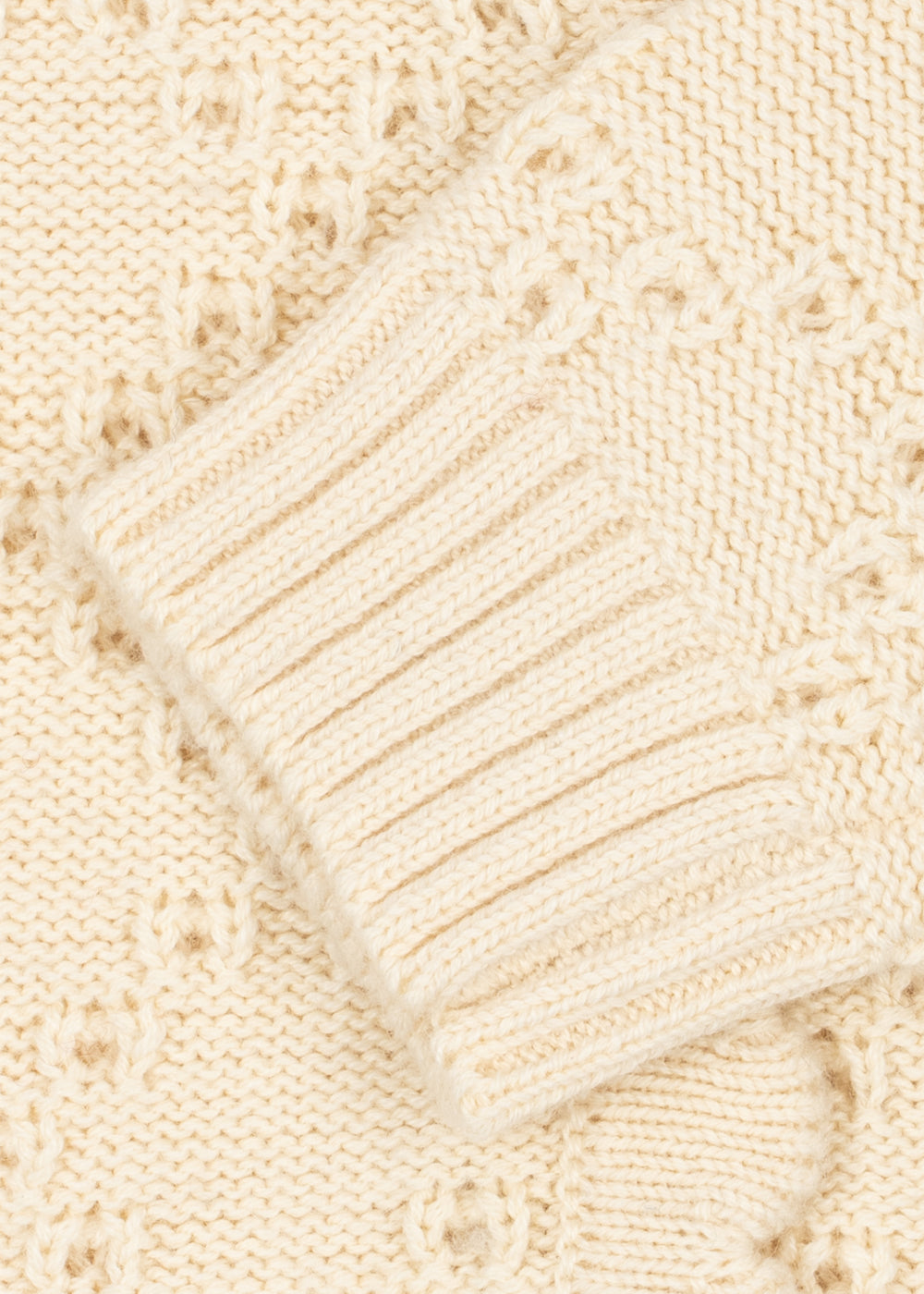 Close-up of a cream-colored knit sweater showing a ribbed cuff and textured openwork knit pattern.