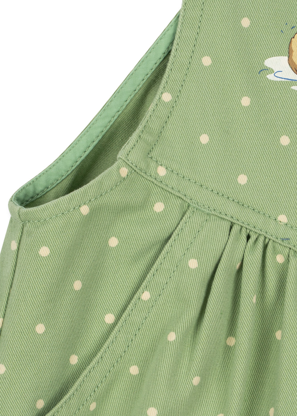 Close-up of a light green children's dress with white polka dots, showing the shoulder strap, neckline, and pleated skirt detail.