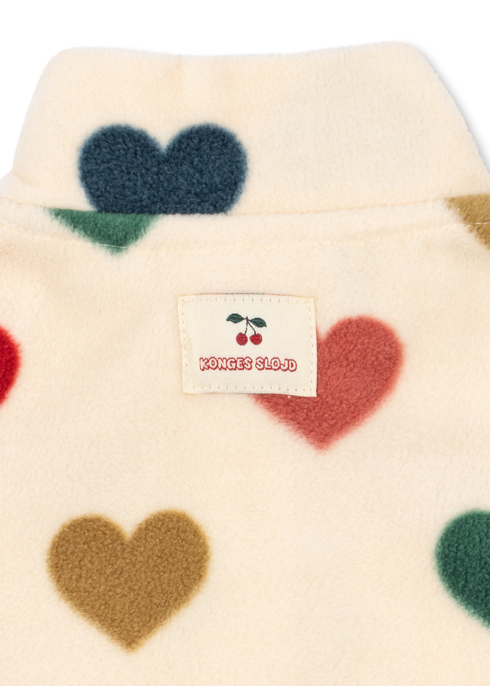 Close-up of a cream-colored fuzzy garment with large heart shapes in navy, red, green, brown, and tan, featuring a sewn-on brand label that reads 'KONNES CLOJ' with a cherry graphic.