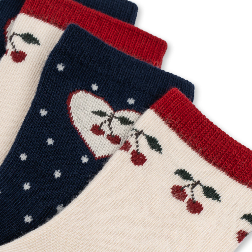 Three pairs of ankle socks arranged diagonally, featuring cherry and heart knit patterns. The socks come in cream, navy blue, and red, with the cream pair in the foreground showing a cherry motif and red cuff, the navy pair in the middle with a heart and white stars, and the top pair partially visible in red and navy.