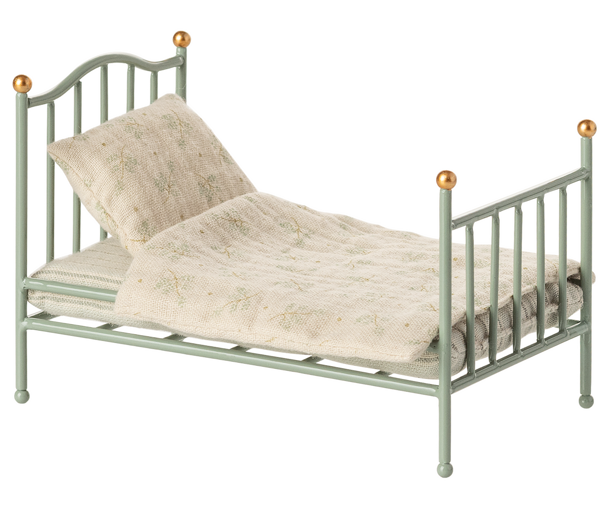 A miniature dollhouse-style metal bed frame in a pale sage green color, styled with a matching quilted cream quilt and pillow featuring a delicate floral pattern, set against a plain white background.